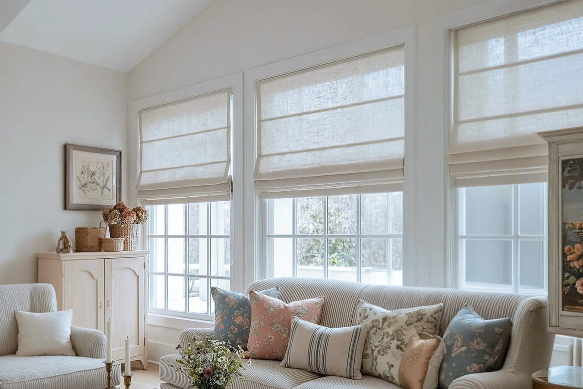 Three beige Roman shades hang above a sofa with patterned cushions in a living room with a sloped ceiling.