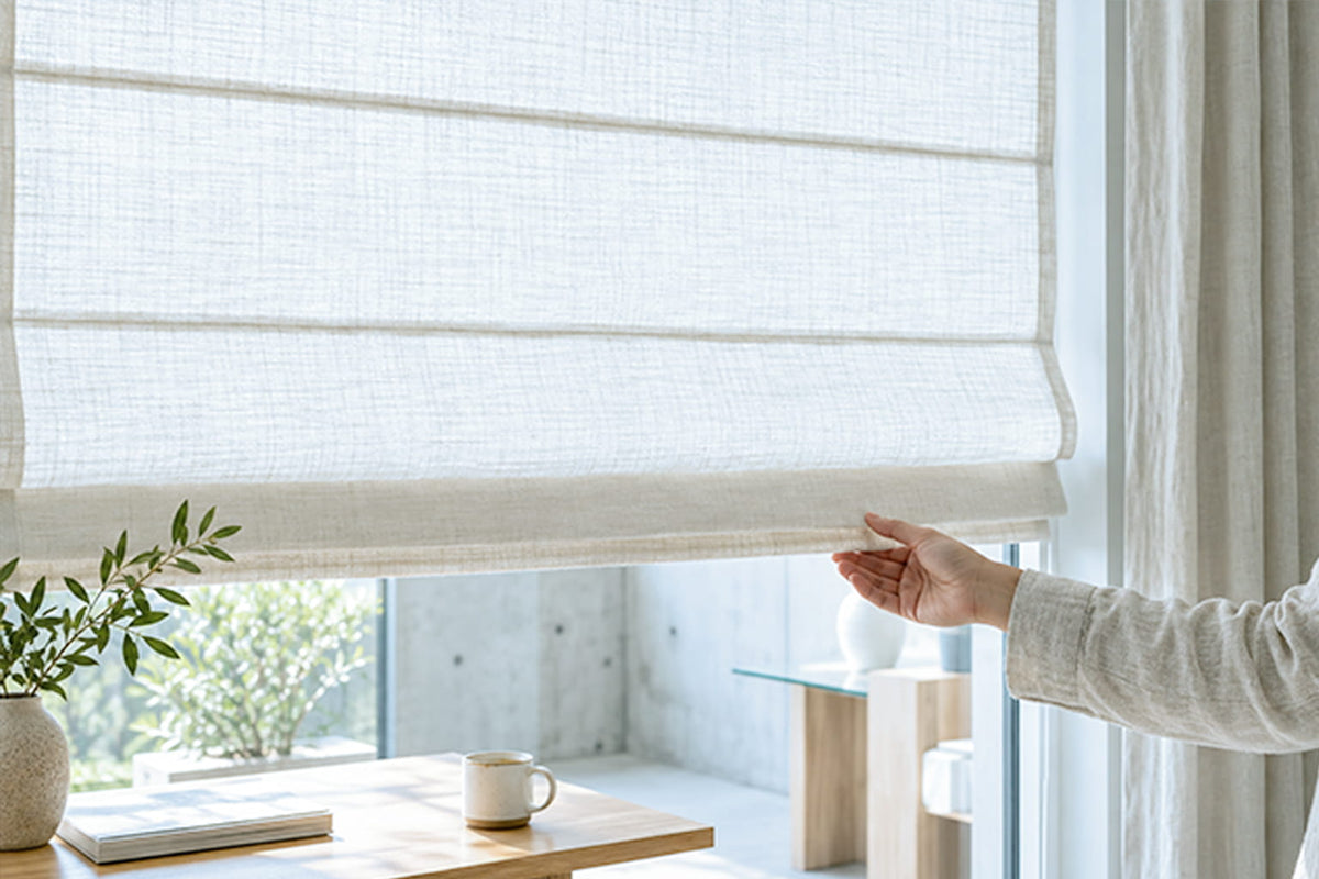 A hand lifts the bottom rail of a white cordless Roman shade above a table with a coffee cup.