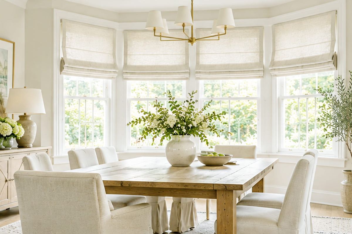 Cream Roman shades follow the angled windows around a dining table with white upholstered chairs.