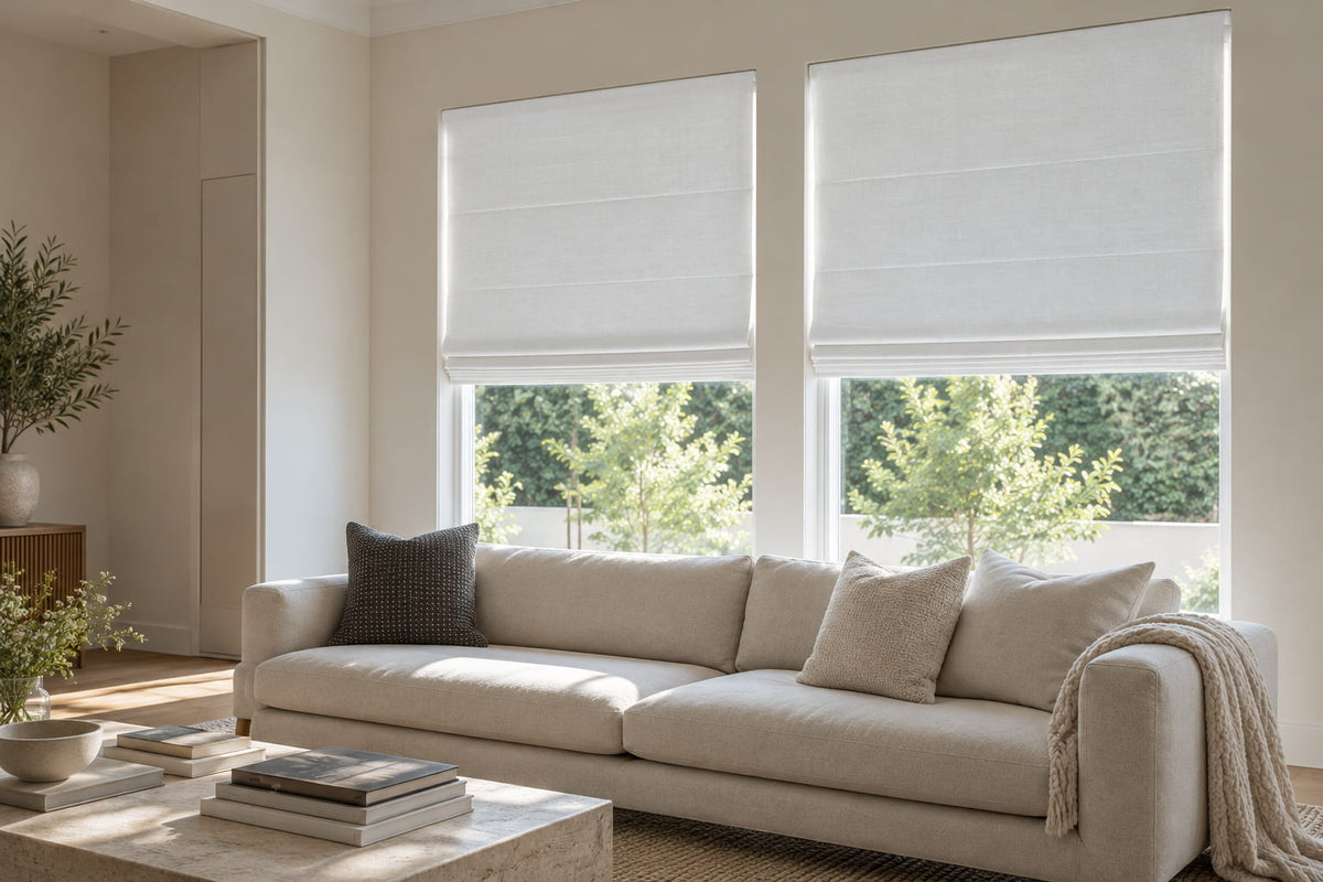 White Roman shades on two living room windows above a cream sofa overlooking leafy trees.