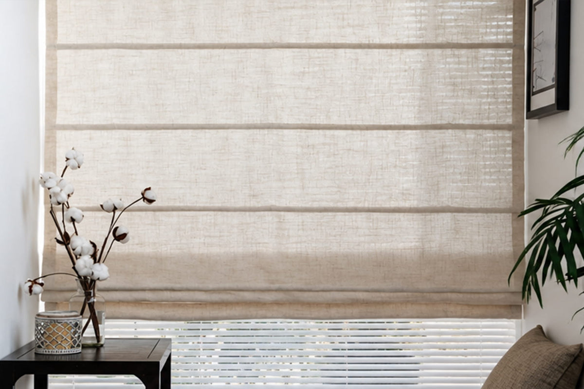 A textured beige Roman shade is lowered across a large window beside a potted orchid and leafy plant.