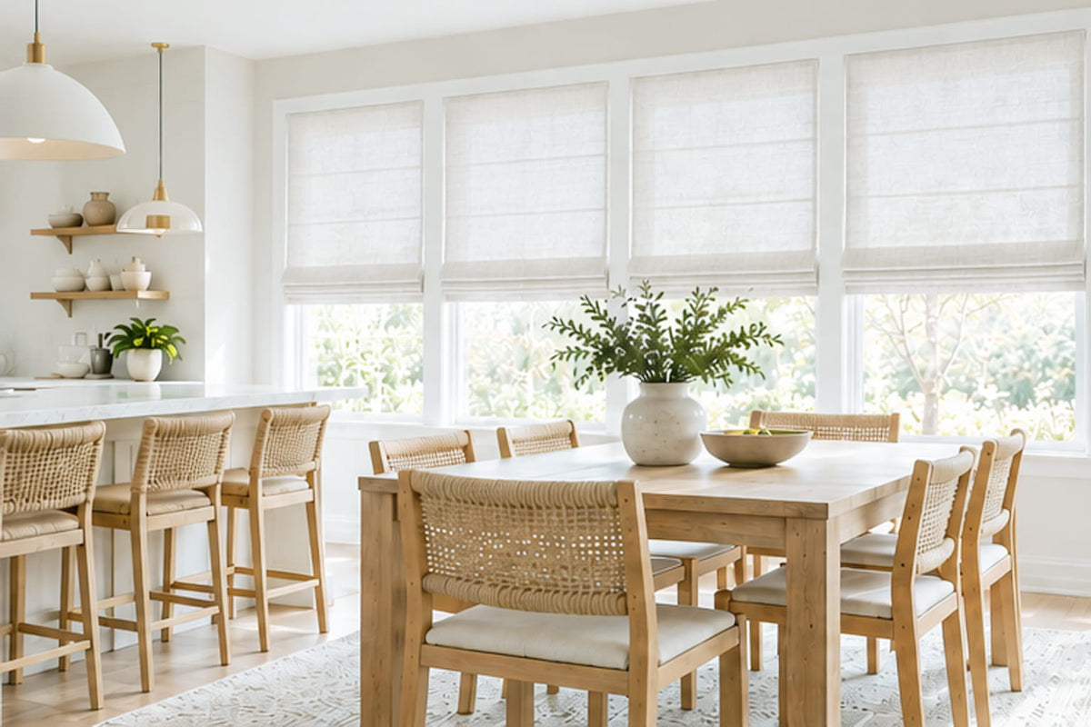 White Roman shades line the windows beside a wooden dining table, woven chairs, and a kitchen breakfast bar.