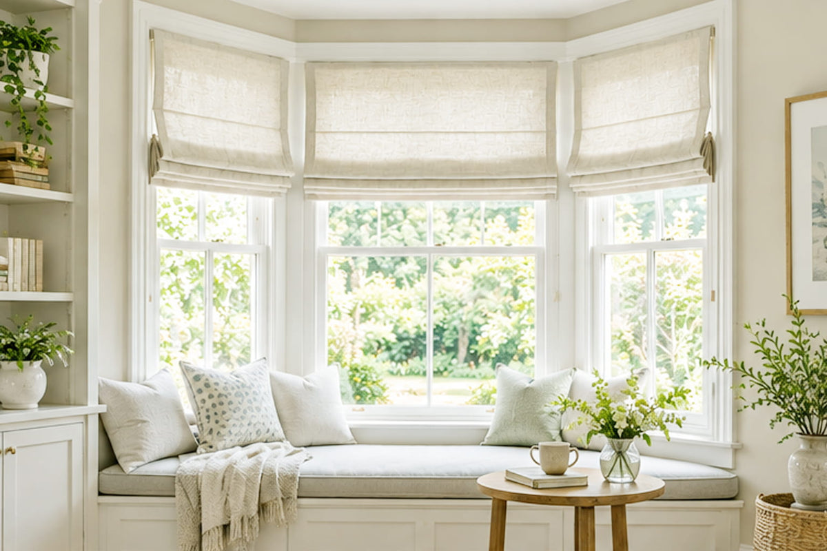 Beige Roman shades hang above a cushioned bay window seat beside bookshelves and a small round table.