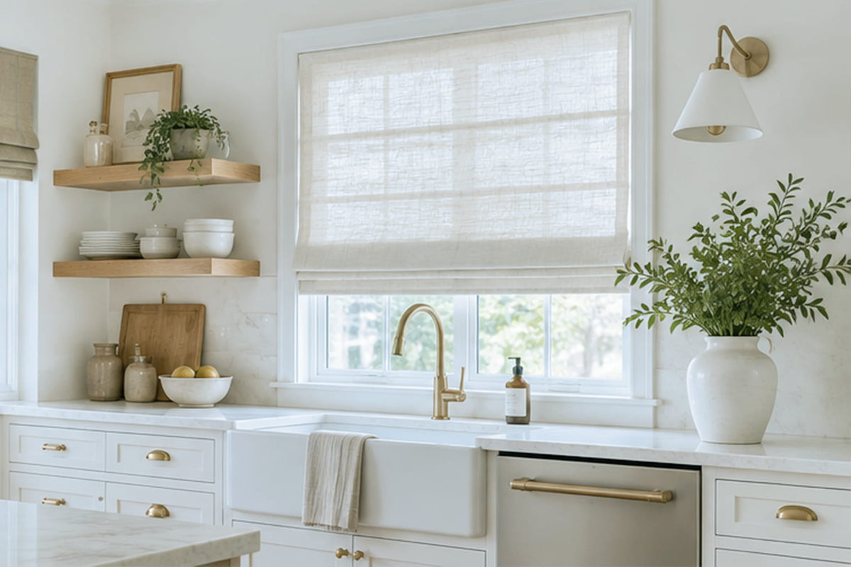 A white Roman shade covers the upper part of a kitchen window above a farmhouse sink and brass faucet.